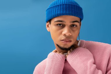 Şık giyinmiş genç adam, parlak mavi bir ortamda düşünceli bir şekilde poz veriyor, modern moda sergiliyor..