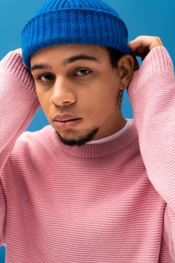 Moda kıyafetli yakışıklı genç adam mavi bir arkaplana karşı güvenle poz veriyor.