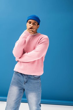 Şık bir genç adam pembe bir süveter ve mavi bir bereyle canlı bir arkaplanda güven içinde poz veriyor..