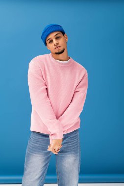 Pembe süveter ve mavi bereli genç bir adam cesur mavi bir arkaplana karşı güvenle poz veriyor..