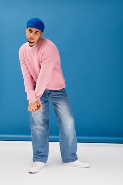 Pembe süveter ve bereli genç bir adam mavi arka planda rahatça poz veriyor..