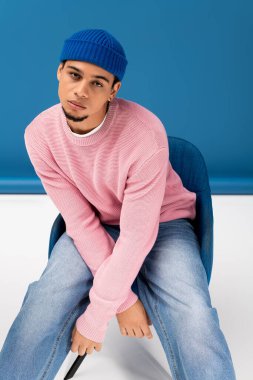 Şık bir genç adam kendine güvenerek poz veriyor, çarpıcı mavi bir arka plana karşı canlı bir kıyafet giyiyor..