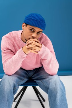 Genç bir adam kendine güvenerek oturur, pembe bir kazak ve mavi bir bere giyer, modern bir tarz yayar..