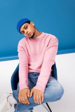 Pembe kazaklı ve mavi bereli yakışıklı genç adam şık bir sandalyede rahatlar, kendine güvenir..