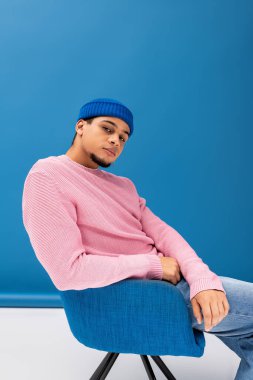 Genç adam mavi bir sandalyede, pembe bir kazak ve mavi bir bere giyerek rahatlıyor..