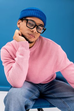Pembe süveter ve mavi bereli şık bir genç adam mavi bir fonda rahatça poz veriyor..