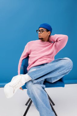 Pembe kazaklı ve mavi şapkalı yakışıklı genç adam bir sandalyede rahatlıyor ve modaya uygun tarzını sergiliyor..