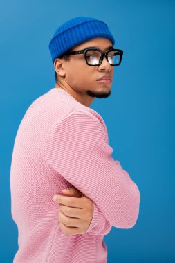 Genç adam, moda görünümünü pembe kazak ve mavi şapkayla sergiliyor..