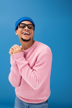 Şık bir genç adam, parlak bir şekilde gülümser. Modaya uygun giysiler içinde, mavi bir arka plana karşı..