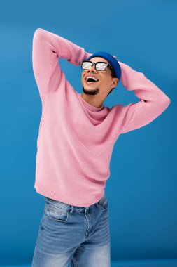 Şık bir genç adam pembe bir kazak ve mavi bir şapka giyer, canlı bir ortamda heyecanla gülümser..