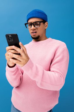 Pembe süveter ve mavi şapkalı genç bir adam telefonunu kontrol ederken şaşırmış görünüyor..