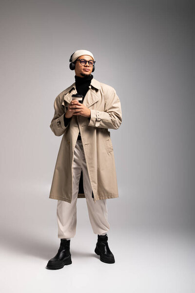 Стильный молодой человек уверенно стоит с напитком, в плаще и наушниках.