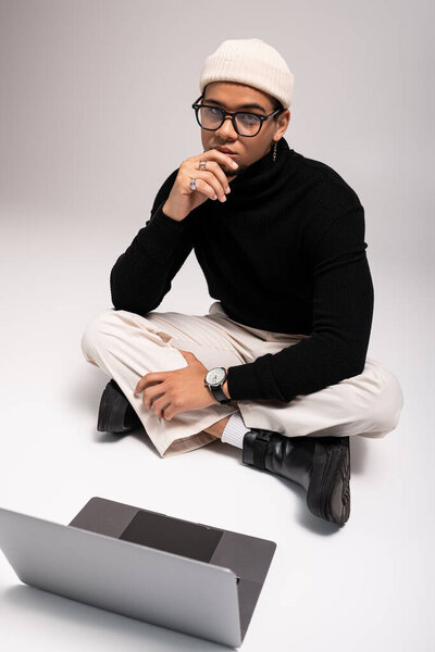 Стильный молодой человек сидит на полу, одетый модно и погруженный в мысли.