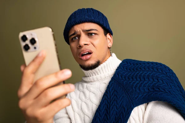 Yakışıklı, şık bir süveter giyen genç bir adam ve beresi telefonuna sürpriz bir şekilde bakıyor..