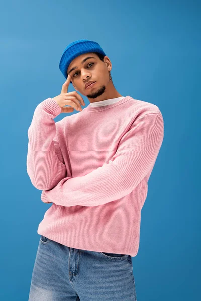 Genç bir adam mavi bir fona karşı pembe bir kazak ve mavi bir şapkayla güvenle poz veriyor..