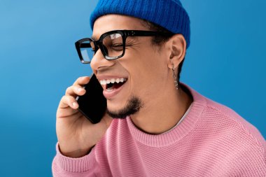 Pembe kazak giyen ve mavi bereli neşeli genç bir adam akıllı telefonuyla konuşuyor, içten bir şekilde gülümsüyor..