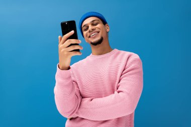 Hoş bir süveter giyen şık genç bir adam mavi bir arka planda akıllı telefonuna gülümsüyor..