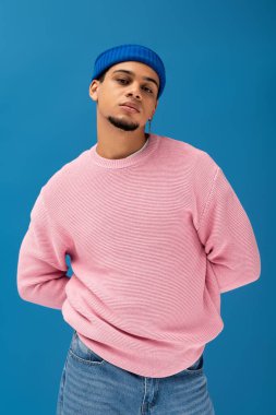 Yakışıklı genç bir adam pembe bir kazak ve mavi bir bere giyer, mavi bir duvara serin bir poz verir..