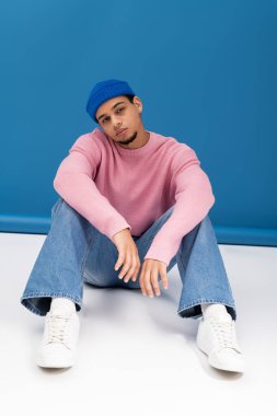 Moda kıyafetleri içindeki genç adam parlak mavi bir arka plana karşı kendine güvenle poz veriyor..