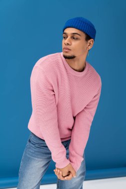 Pembe kazaklı ve mavi bereli kendinden emin genç adam parlak bir arka plana karşı poz veriyor..