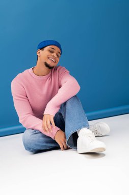 Şık bir genç adam neşeyle mavi bir arka plana yaslanır, büyüleyici ve kendine güvenlidir..