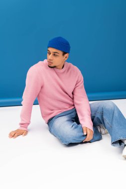 Pembe süveter ve mavi bereyle yakışıklı genç adam yerde oturmuş, moda gösterisi yapıyor..