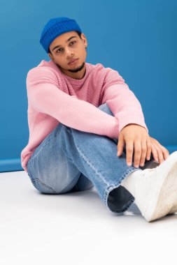 Şık giyinmiş genç adam rahat bir şekilde oturur, canlı bir arkaplana karşı güven yayar..