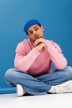 Pembe kazak ve kot pantolon giymiş yakışıklı genç bir adam canlı mavi arka planda oturuyor..