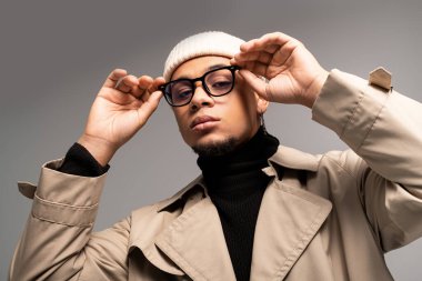 Modaya uygun trençkotlu ve gözlüklü genç adam stüdyoda modern tarz ve karizmayı ifade ediyor..