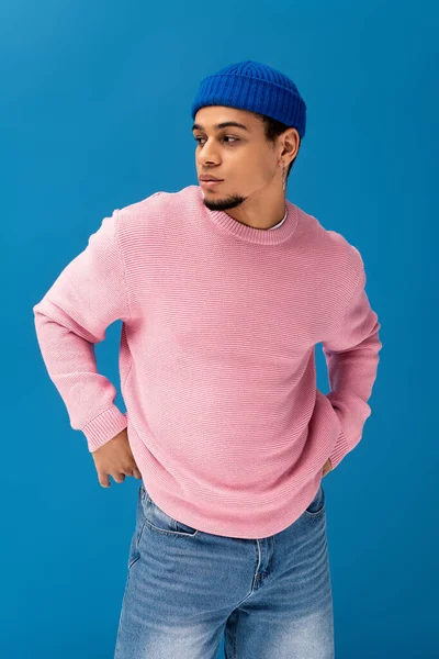 Yakışıklı genç adam kendine güvenerek duruyor, gösterişli pembe süveterini ve mavi şapkasını gösteriyor..