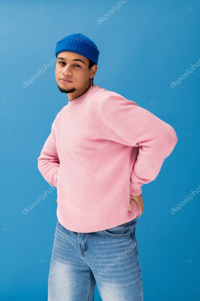 Stylish young man poses confidently in a pink sweater and blue beanie with blue backdrop.