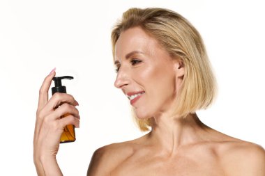 Güzel sarışın bir kadın elinde bir cilt bakım ürünü tutarken gülümsüyor..