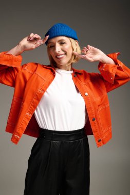 Şık, sarışın bir kadın stüdyoda poz verirken parlak turuncu bir ceket ve mavi bir bere giyiyor..