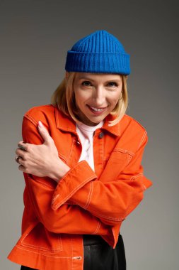 Turuncu ceketli ve mavi bereli sarışın bir kadın stüdyo odasında sıcacık poz veriyor..