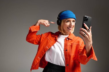 Mavi bereli ve turuncu ceketli modaya uygun sarışın bir kadın stüdyoda neşeyle selfie çekiyor..