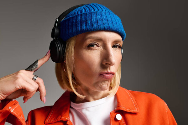 Стильная женщина с короткими светлыми волосами уверенно позирует в голубой шапочке и оранжевой куртке.