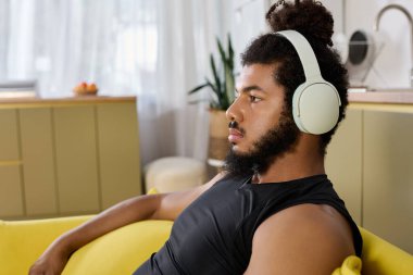 Sakallı Afrikalı Amerikalı bir adam sarı bir kanepede dinleniyor ve müzik dinliyor.