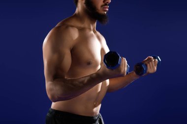 Sakallı ve kıvırcık saçlı atletik bir adam, çarpıcı mavi bir alanda ağırlık kaldırıyor..