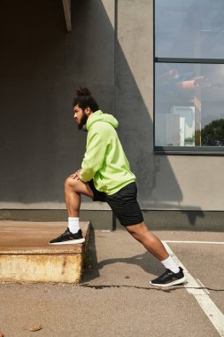 Yeşil kapüşonlu bir adam dışarıda bankta bacağını esnetiyor. Egzersiz yapıyor..