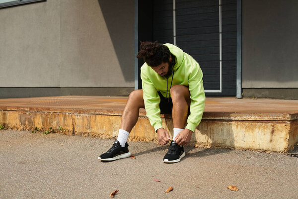 A man prepares for his workout outside, tying his shoes while wearing a bright green hoodie.