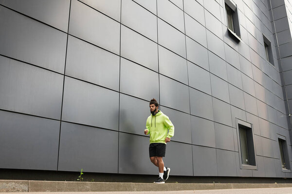 An athletic African American man works out by jogging outdoors, dressed in a green hoodie.