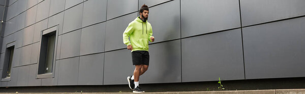 A young african american man jogs along a modern building's wall wearing a green hoodie and shorts, banner