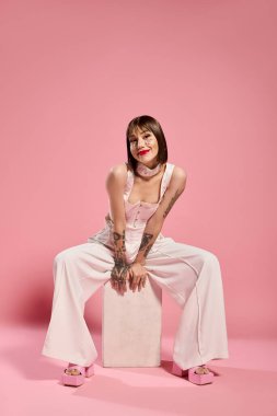 Şık giyinen genç bir kadın pembe bir stüdyoda otururken güzelliğini sergiliyor..