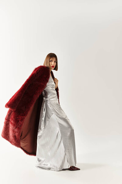 Chic young woman poses dramatically in a shiny silver gown, embellished by a vibrant red coat.