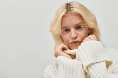 Beyaz kirpikli genç sarışın bir kadın tarafsız bir arkaplan karşısında rahat bir süveter mankenliği yapıyor..