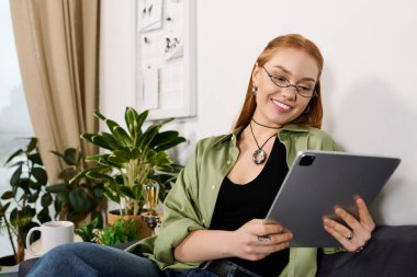 Güzel genç bir kadın yeşil bitkilerin arasında tablet kullanırken gülümseyerek evinde dinleniyor..