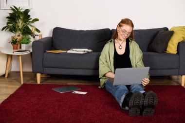 Genç bir kadın rahat bir şekilde kırmızı halıda oturuyor. Dizüstü bilgisayarıyla rahat bir evde oturuyor..