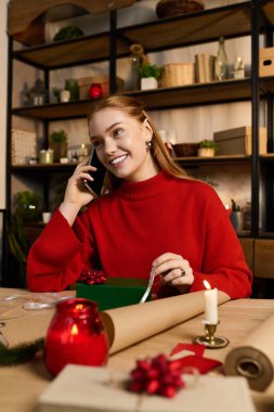 Kırmızı kazaklı genç bir kadın, rahat evinde hediyelerini paketlerken bir telefon görüşmesi yapmaktan hoşlanıyor..