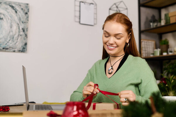 Молодая женщина улыбается и аккуратно обвязывает ленту вокруг красиво обернутого подарка дома.