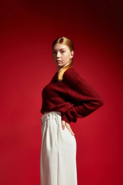 Kızıl saçlı şık bir genç kadın cesur kırmızı bir arka fonun önünde rahat bir süveter sergiliyor..